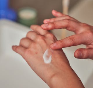 A person applies a small amount of hydroquinone cream to the back of their hand near a sink area.