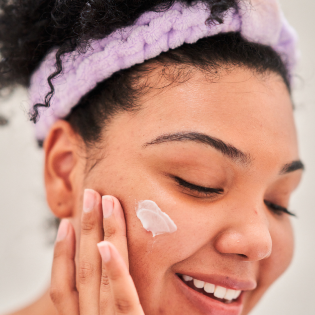 A person applies hydroquinone cream to their cheek while smiling, wearing a purple headband.