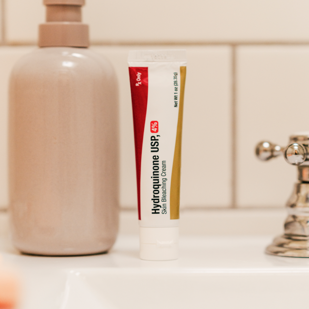 A tube of Hydroquinone Cream USP 2% sits on the bathroom sink beside a soap dispenser, framed by a tiled wall in the background.