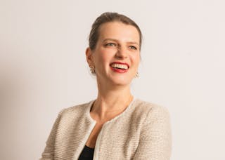 A person with tied-back hair and a light-colored jacket smiles against a plain background.