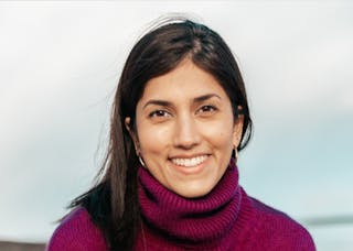A person with long dark hair smiles at the camera, wearing a purple turtleneck sweater. The background is a blurred sky.