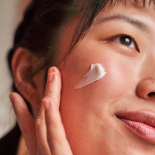 A person applies a dab of tretinoin cream to their cheek, smiling softly.