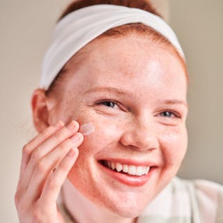 A person with a headband applies Tretinoin Cream to their cheek, smiling.