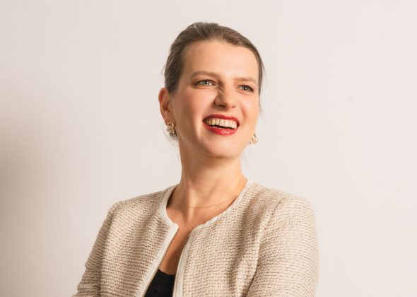 A person with tied-back hair and a light-colored jacket smiles against a plain background.