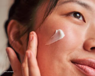 Close-up of a person applying a small dab of cream to their cheek with fingers, smiling slightly.