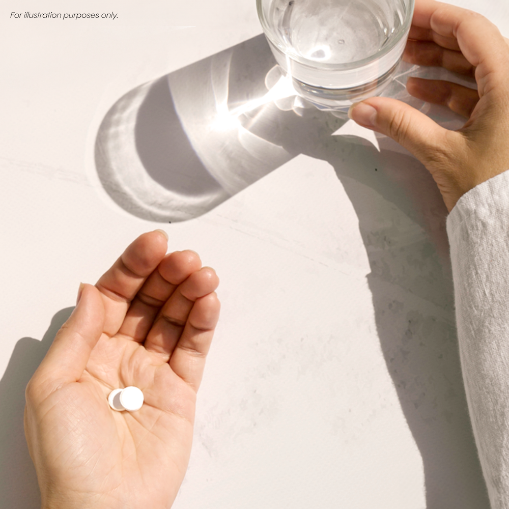 A person holds two white pills in one hand and a glass of water in the other on a white surface.