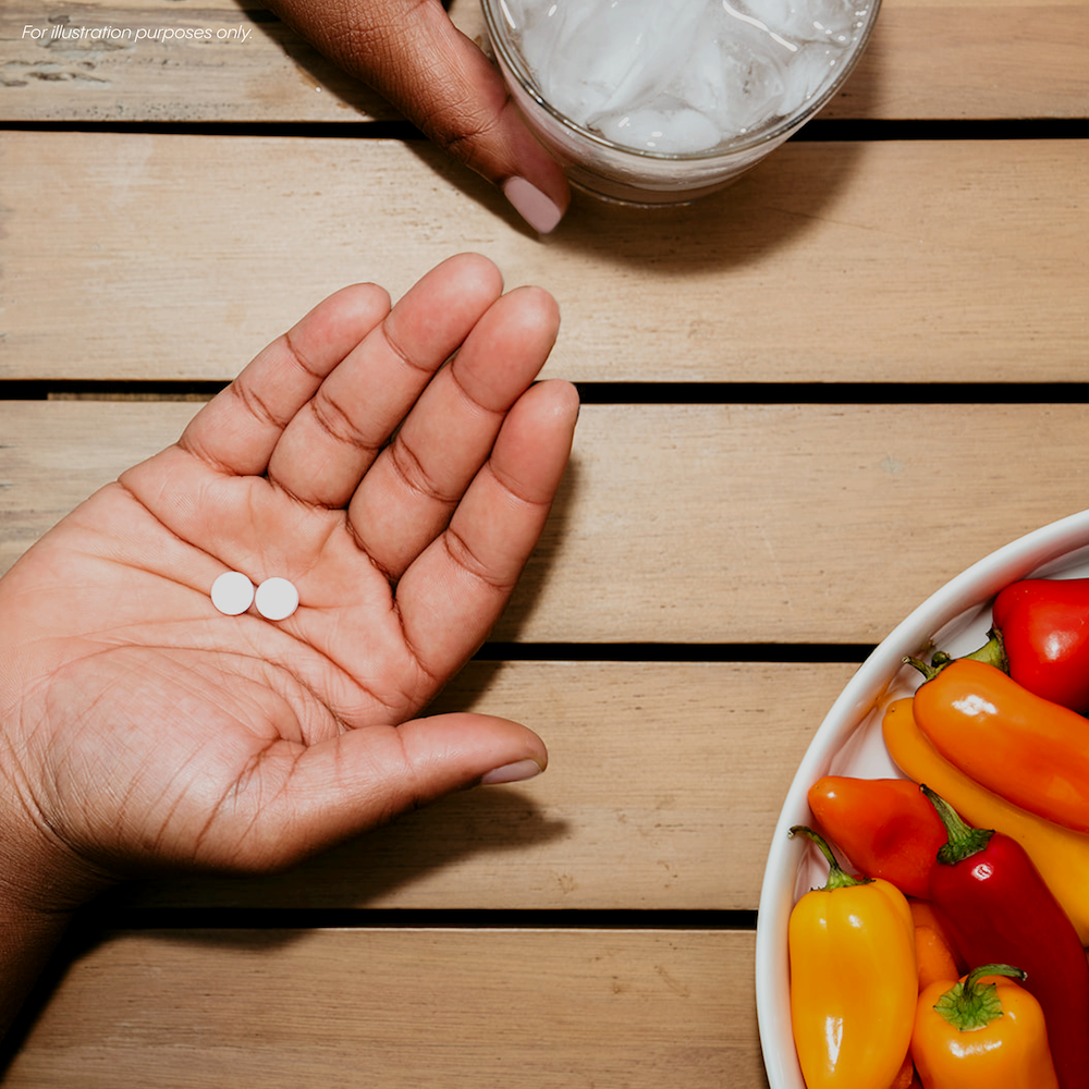 Hand holding two white pills above a bowl of peppers on a wooden table, with another hand holding a glass of ice water nearby.