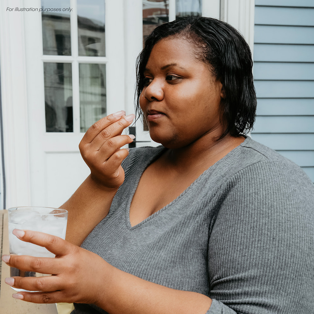 A person in a gray shirt holds a glass of ice water and a pill near their mouth in front of a blue house exterior.