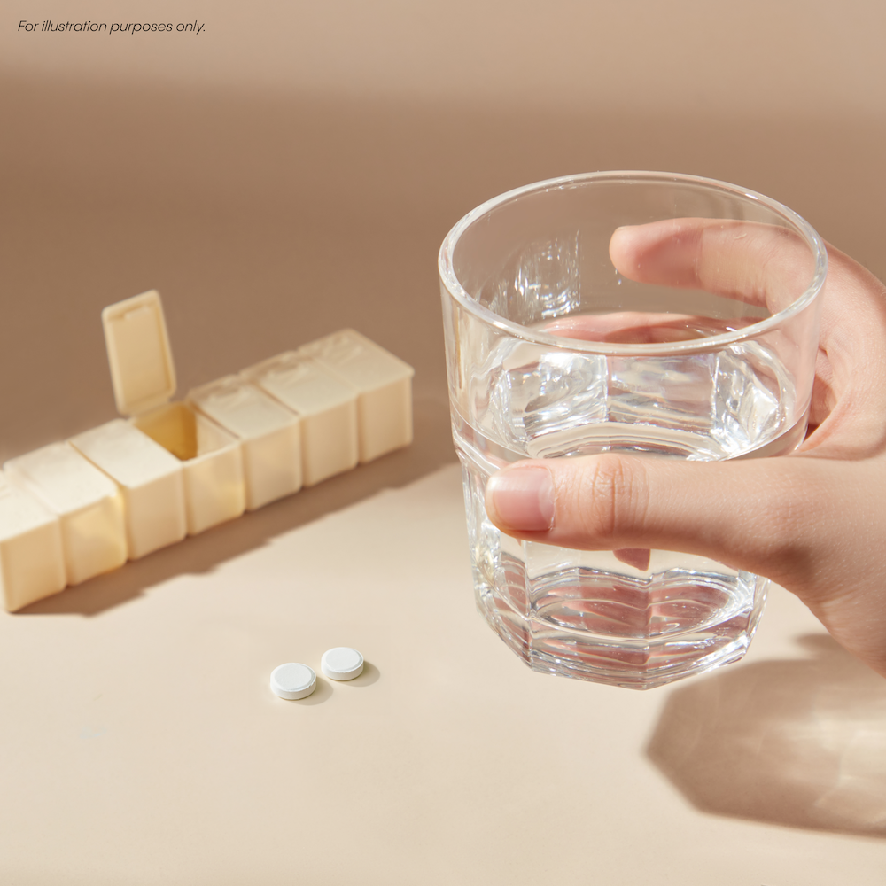 A hand holds a glass of water near a pill organizer. Two white tablets are on the table.