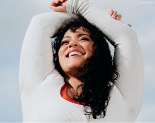 Person stretching outdoors, wearing a white long-sleeve shirt, smiling with arms raised overhead against a blue sky background.