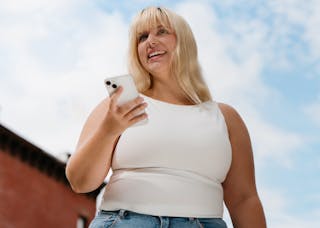A person with long blond hair holds a smartphone outdoors, smiling and looking up. They're wearing a white sleeveless top and jeans against a background of a blue sky and a brick building.