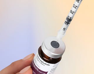 A hand holds a vial labeled as a serum, while a syringe is inserted into the rubber stopper on top.