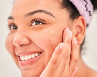 Person applying skincare product to their cheek, smiling, with a focus on their face and a headband holding back their hair.