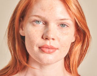 A woman with fair skin, red hair, and freckles looks directly at the camera with a neutral expression against a light background.
