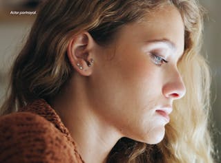 A woman with blonde hair and multiple ear piercings looks down pensively, wearing a brown sweater. Text in the corner reads 