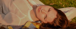 A person with long brown hair lies on a blanket outdoors, looking up towards the camera, with sunlight illuminating their face.