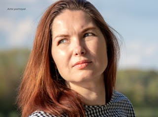 A woman with long auburn hair wearing a black and white checkered top looks slightly upward outdoors on a sunny day.