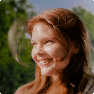 A young woman with red hair smiles outdoors, with sunlight on her face and greenery blurred in the background.